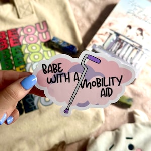 Könnte beinhalten: Ein Aufkleber mit dem Text "Babe with a Mobility Aid" in schwarzer Schrift, der ein Cartoon-Bild eines Gehstocks zeigt. Der Hintergrund ist eine rosa und lila Wolkenform. Der Aufkleber wird in einer Hand mit blauem Nagellack gehalten.