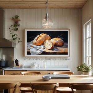 May include: A framed painting of three loaves of freshly baked bread on a white linen cloth. The bread is golden brown and has a rustic appearance. The painting is set against a dark background and is displayed in a modern kitchen.