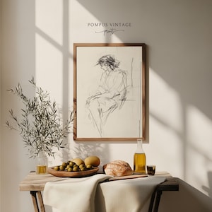 May include: A framed black and white line drawing of a woman sitting in a chair. The drawing is on a cream background and is framed in a natural wood frame. The frame is hanging on a white wall. There is a wooden table with a bowl of olives, a loaf of bread, a bottle of olive oil, and a glass on it. The table is covered with a white tablecloth.