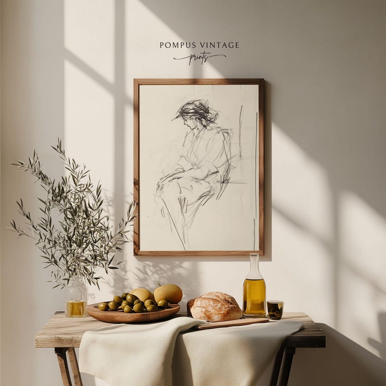 May include: A framed black and white line drawing of a woman sitting in a chair. The drawing is on a cream background and is framed in a natural wood frame. The frame is hanging on a white wall. There is a wooden table with a bowl of olives, a loaf of bread, a bottle of olive oil, and a glass on it. The table is covered with a white tablecloth.