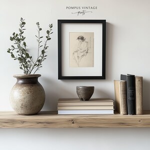 May include: A black framed print of a pencil drawing of a person sitting on a chair. The print is on a shelf with a vase of greenery, a small bowl, and a stack of books.