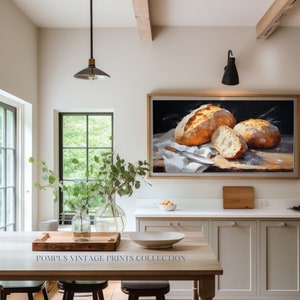 May include: A framed painting of three loaves of freshly baked bread on a white linen cloth. The painting is set against a dark background and features a knife on the cloth. The painting is hanging on a white wall in a kitchen setting.