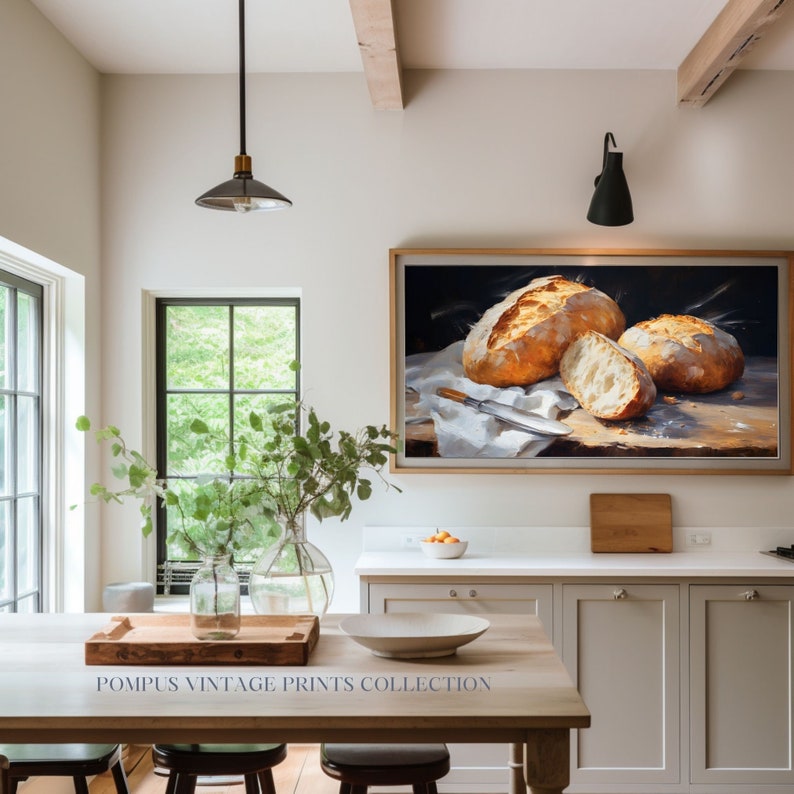 May include: A framed painting of three loaves of freshly baked bread on a white linen cloth. The painting is set against a dark background and features a knife on the cloth. The painting is hanging on a white wall in a kitchen setting.