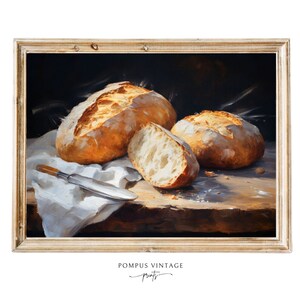 May include: A painting of three loaves of bread on a wooden table with a white cloth. The bread is golden brown and crusty. The painting is framed in a light wood frame.