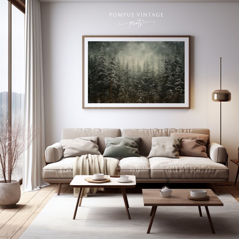May include: A framed print of a misty forest scene with tall evergreen trees. The print is hanging on a white wall above a beige sofa with throw pillows and a blanket. The sofa is in a living room with a wooden floor and a coffee table.