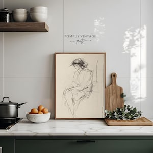 May include: A framed black and white line drawing of a woman sitting in a chair. The drawing is on a cream background and the frame is a light brown wood. The drawing is titled "Pompous Vintage Prints".