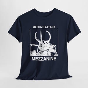 May include: Navy blue t-shirt featuring a white graphic of a stylized insect with the text "MASSIVE ATTACK" above and "MEZZANINE" below. The design is enclosed in a white square. The shirt has short sleeves and a crew neck.