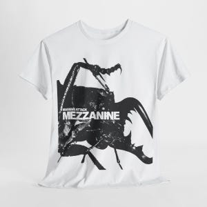 May include: White t-shirt featuring a large black and white graphic of an insect with the text "MASSIVE ATTACK MEZZANINE" in a bold, sans-serif font. The shirt has short sleeves and a crew neck.