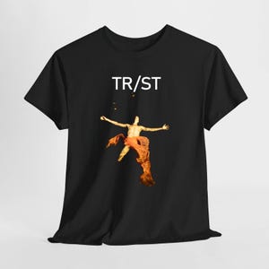 Puede incluir: Una camiseta negra de manga corta con el texto blanco TR/ST centrado sobre un gráfico de una figura con los brazos extendidos, envuelta en una tela naranja fluida sobre un fondo oscuro. El diseño está impreso en la parte frontal de la prenda.