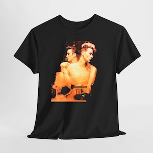 Puede incluir: Una camiseta negra de manga corta con un estampado gráfico central en tonos naranja y negro. El diseño muestra a dos figuras posicionadas espalda contra espalda con un efecto texturizado y desgastado, y el texto "IN THE MOONLIGHT" debajo.