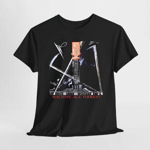 Puede incluir: Una camiseta negra de manga corta con un estampado gráfico de una torre alta y estilizada con líneas geométricas blancas. El diseño incluye un emblema rectangular naranja en la parte superior y el texto rojo MACHINE AGE VOODOO abajo.