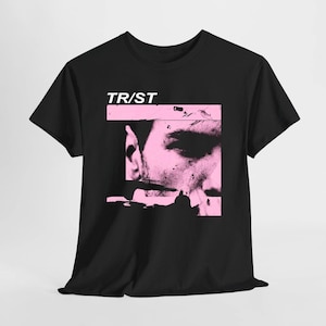 Puede incluir: Una camiseta negra de manga corta con un estampado gráfico en un marco rectangular de color rosa claro. El estampado muestra un primer plano en blanco y negro de alto contraste de un rostro con el texto TR/ST impreso en blanco.
