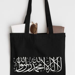 Könnte beinhalten: Schwarze Tragetasche mit weißer arabischer Kalligraphie. Die Tasche hat zwei schwarze Griffe und enthält ein weißes Buch, eine Sonnenbrille und eine braune Brieftasche. Die Kalligraphie ist das Hauptdesign.