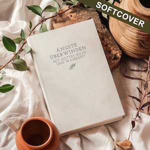May include: A cream-coloured softcover book with the title "ÄNGSTE ÜBERWINDEN" in dark green lettering. The book is open on a white surface with text that reads "MIT GOTTES HILFE UND WAHRHEIT". The background includes greenery and a small terracotta pot.
