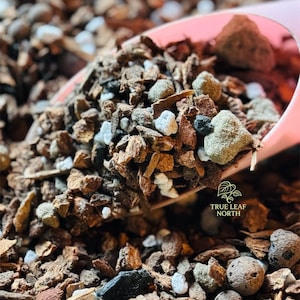 May include: A pink scoop holds a blend of soil components: brown bark, white and grey pebbles, and dark charcoal. The image shows a close-up of the mix, with the True Leaf North logo in the bottom right, indicating a gardening product.