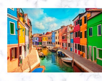 Può includere: Un canale stretto fiancheggiato da case colorate in un villaggio europeo. Le case sono dipinte in tonalità di blu, giallo, verde, rosso e arancione. Piccole barche sono ormeggiate lungo il canale.