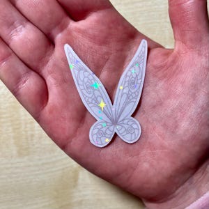 May include: A white iridescent sticker featuring a detailed butterfly design. The butterfly has a white outline and is filled with iridescent glitter.