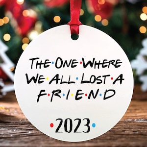 Op de afbeelding: Ronde witte kerstbal met de tekst "The One Where We All Lost A Friend" en het jaar "2023" in zwarte tekst met gekleurde stippen.