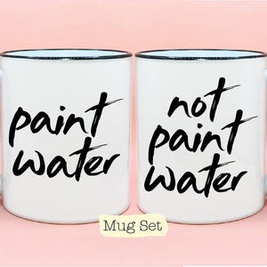 May include: A set of two white ceramic mugs with black handles. One mug says "paint water" and the other mug says "not paint water".