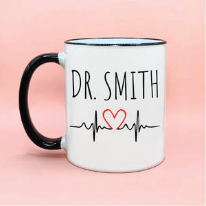 以下が含まれることがあります： 黒いハンドルと縁が付いた白いセラミックマグ。マグには、黒で「DR. SMITH」という文字、赤いハート、心拍ラインのデザインが施されています。マグは標準サイズです。