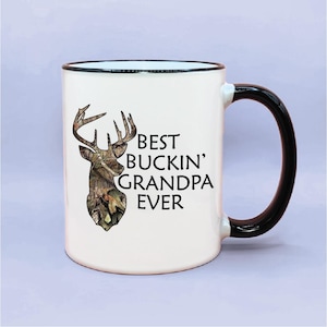 May include: White ceramic coffee mug with a black rim and handle. The mug features a camouflage deer head with antlers and the text "BEST BUCKIN' GRANDPA EVER".