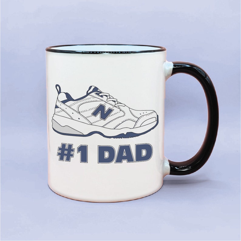 May include: White ceramic mug with a black rim and handle. The mug features a black and white illustration of a running shoe with the text "#1 DAD" in blue.