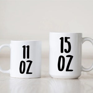 May include: Two white ceramic mugs with black text. The smaller mug says "11 oz" and the larger mug says "15 oz".