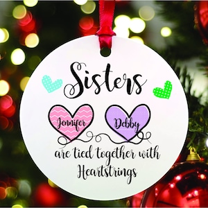 May include: White round Christmas ornament with the text "Sisters are tied together with heartstrings". Two pink and purple hearts with the names "Jennifer" and "Debby" are on the ornament.