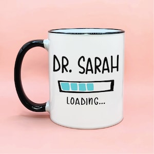 May include: White ceramic mug with a black handle and rim. The mug has black text that reads "DR. SARAH" and a blue progress bar that says "LOADING..." underneath.