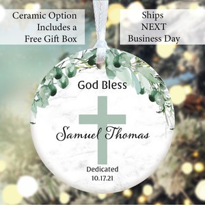 May include: A white ceramic ornament with a green cross and the text "God Bless Samuel Thomas" and "Dedicated 10.17.21". The ornament is decorated with green watercolor leaves and a marble-like background.