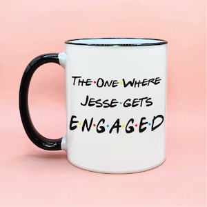 Puede incluir: Taza de cerámica blanca con asa y borde negros. La taza tiene un gráfico negro que dice "The One Where Jesse Gets Engaged" con puntos de colores.
