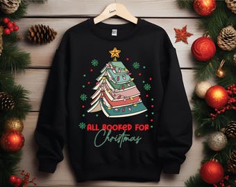 May include: Black sweatshirt with a Christmas tree design made of books, adorned with a star and string lights. The text "ALL BOOKED FOR Christmas" is written in green and white. Festive holiday decor surrounds the sweatshirt.