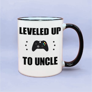 Może przedstawiać: Biały kubek ceramiczny z czarnym rantem i uchwytem. Kubek ma czarny napis "LEVELED UP TO UNCLE" z czarnym obrazkiem kontrolera do gier wideo w środku.