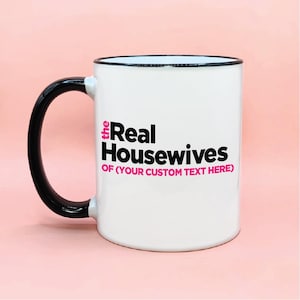 May include: White ceramic mug with a black handle and rim. The mug features the text "the Real Housewives" in black and pink, with "OF (YOUR CUSTOM TEXT HERE)" below. The mug is set against a pink background.