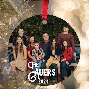 May include: A round Christmas ornament featuring a family portrait. The ornament is white with black text that reads "The Auers 2024". The family is dressed in casual attire and is standing in front of a green background.