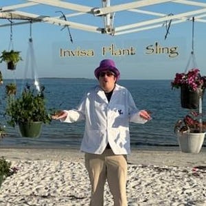 May include: A white canopy frame on a beach holds multiple hanging plant pots suspended by clear straps. The pots contain various green plants and colorful flowers. A sign hanging from the frame reads Invisa Plant Sling in gold lettering.