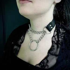 Op de afbeelding: Een zwarte leren choker met een zilveren ketting en een grote zilveren ring. De choker heeft twee zilveren studs aan de achterkant.