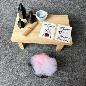 Könnte beinhalten: Miniatur-Hexen-Zaubertrank-Set auf einem kleinen Holztisch. Enthält schwarze Flaschen, einen Mörser und Stößel, eine Schriftrolle und zwei Karten mit Text und Illustrationen. Ein schwarzer Kessel enthält rosa und weißes flauschiges Material.