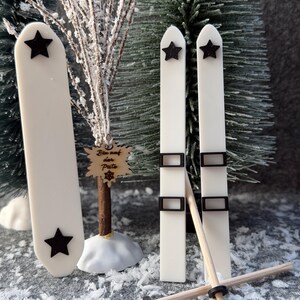 Könnte beinhalten: Drei weiße Deko-Skier mit schwarzen Sternen und schwarzen rechteckigen Bindungen. Zwei Skier stehen aufrecht, einer ist links platziert. Zwei dünne Holzstöcke mit schwarzen Ringen sind vor den Skiern gekreuzt angeordnet.