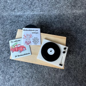 Könnte beinhalten: Miniatur-Plattenspieler in Weiß mit einer schwarzen Schallplatte und einem Holzsockel. Auf dem Sockel liegen zwei kleine Karten, eine mit einem roten LKW und dem Text "24 Weihnachtslieder", die andere mit einem Rentier und dem Text "Weihnachtslieder zum mitsingen".