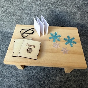 Könnte beinhalten: Ein Miniatur-Holztisch mit zwei quadratischen Holzplatten, die mit dem Wort Presse und einem Schneeflockensymbol graviert sind, einem schwarzen Gummiband, einer gefalteten weißen Papierharmonika und vier transparenten blauen und klaren Schneeflocken.