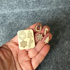 Könnte beinhalten: Eine Sammlung kleiner, unfertiger Weihnachtsornamente aus Holz. Das Set enthält Formen von Kranz, Schneeflocke, Zuckerstange und Weihnachtsbaum. Die Ornamente werden in einer Hand vor einem grauen Hintergrund gehalten.