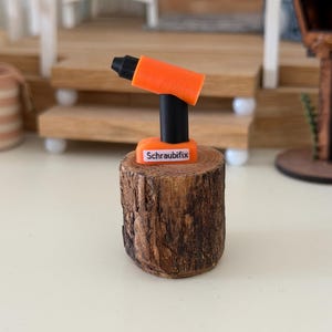 Könnte beinhalten: Ein orange-schwarzer Miniatur-Akkuschrauber mit dem Schriftzug Schraubifix auf der Basis. Das Werkzeug steht aufrecht auf einem kleinen, zylindrischen, strukturierten braunen Holzstamm, der eine realistische Maserung und Rindenstruktur aufweist.