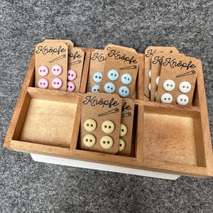 Könnte beinhalten: Sortiment an Knöpfen in einem Holzdisplay. Die Knöpfe sind in vier Farben: rosa, blau, weiß und gelb. Jede Farbe ist mit dem Wort "Knöpfe" beschriftet.