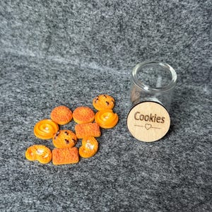 Könnte beinhalten: Eine Sammlung von Miniatur-Keksen in Orange mit verschiedenen Formen, darunter runde, rechteckige und spiralförmige Designs. Ein klares Glas und ein Holzdeckel mit dem Wort "Cookies" und einem Herz sind ebenfalls vorhanden.