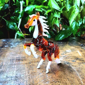 May include: A brown and white glass horse figurine with a white mane and tail. The figurine is standing on a wooden surface.
