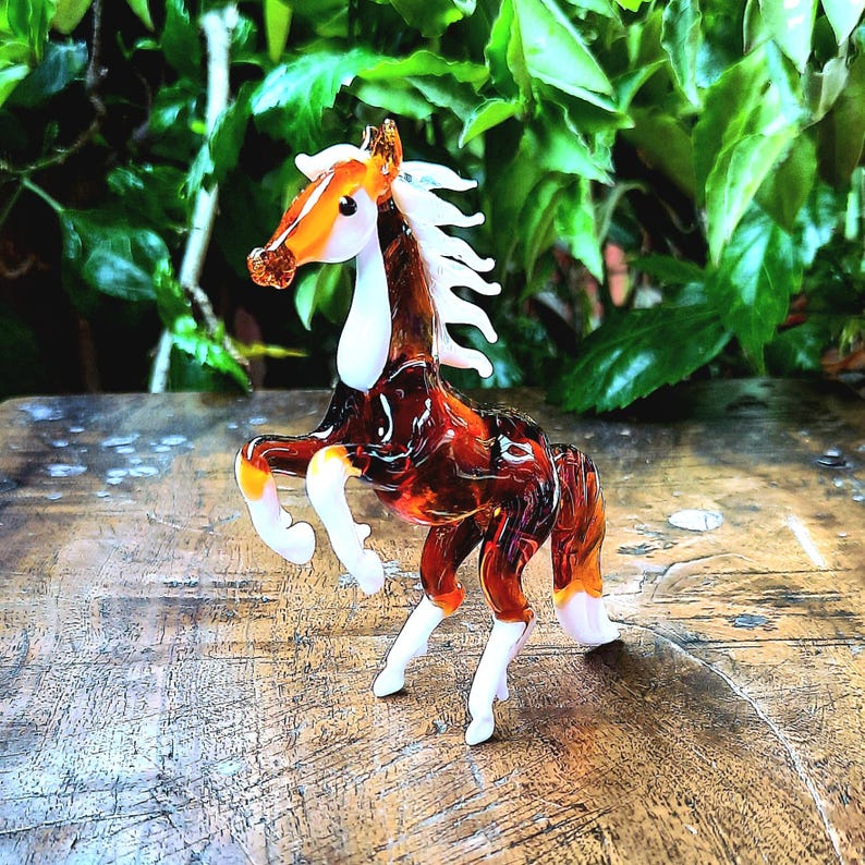 May include: A brown and white glass horse figurine with a white mane and tail. The figurine is standing on a wooden surface.
