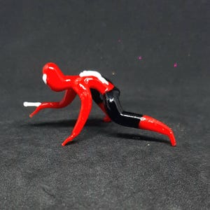 May include: A small, stylized glass figurine in a crawling pose. The figure is primarily red with black and white accents. The legs and arms are elongated, and the figure appears to be in motion. The background is a dark gray.