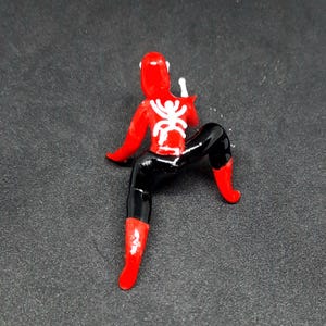 May include: A red and black figurine of a superhero in a crouching pose. The figure has a white spider emblem on the back and is wearing a red suit with black pants and boots. The figurine is on a dark gray surface.