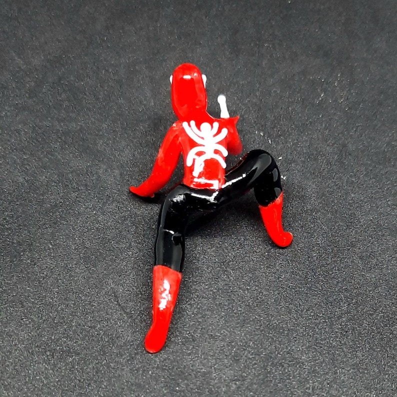May include: A red and black figurine of a superhero in a crouching pose. The figure has a white spider emblem on the back and is wearing a red suit with black pants and boots. The figurine is on a dark gray surface.